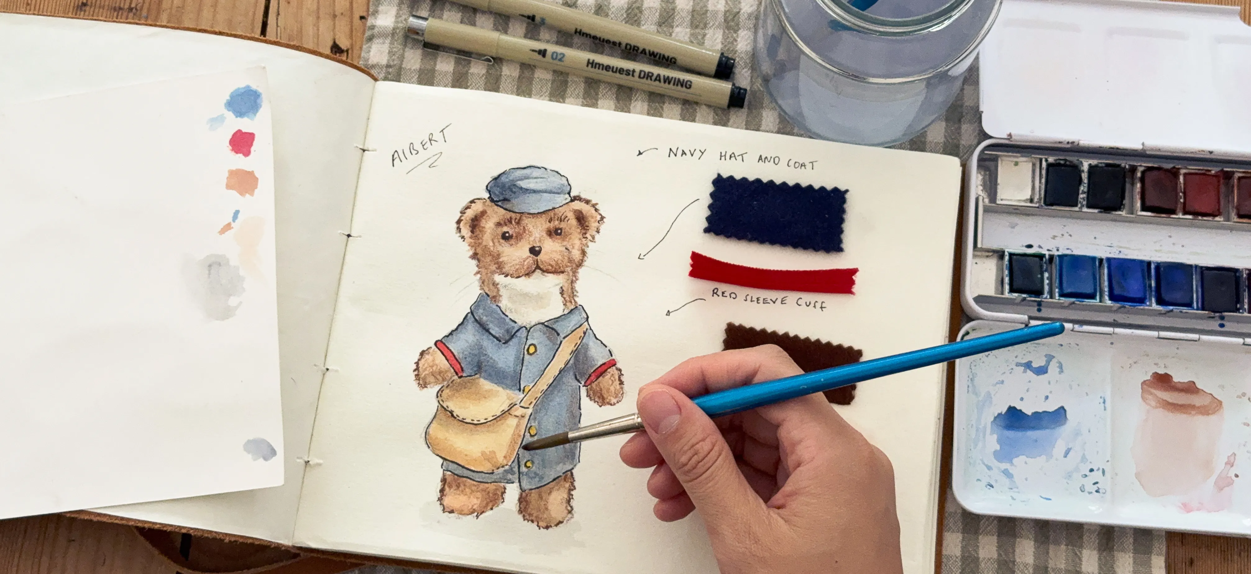 Albert the Postman being hand-painted in a sketchbook, surrounded by watercolours and fabric swatches
