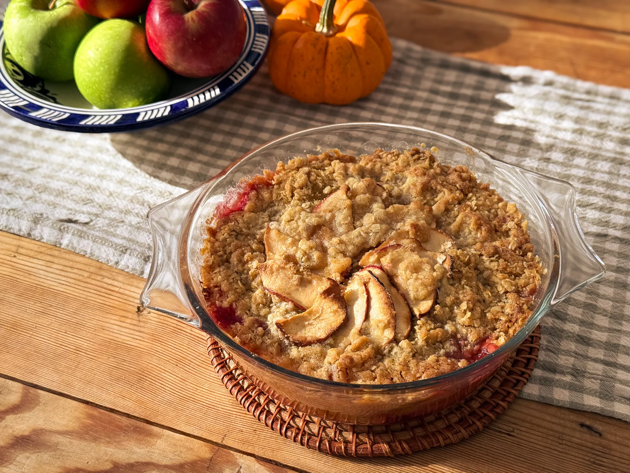 A golden apple and blackberry crumble in a glass dish, surrounded by apples and a small pumpkin
